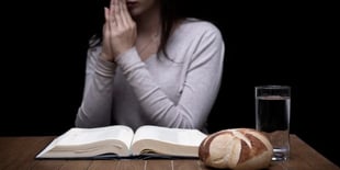 A woman prays near an open Bible, glass of water, and loaf of bread